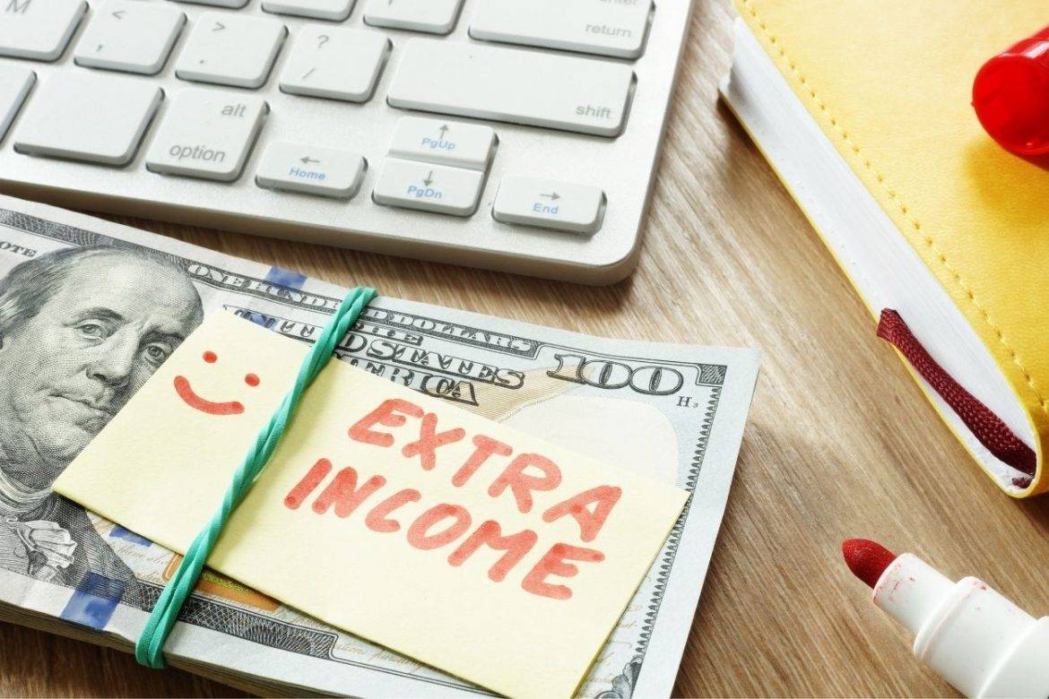 Stack of cash next to red marker and keyboard with note reading Extra Income, indicating person is earning extra income while working full time