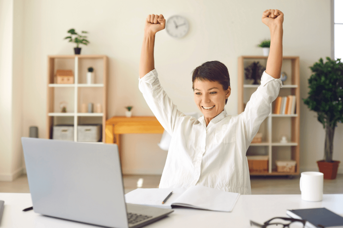 Woman sitting in front of laptop with arms raised in celebration for earning large profit margin by selling promotional products