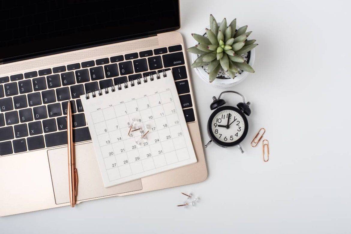 Plant and small clock next to laptop computer with small calendar, pen, and push pins on it