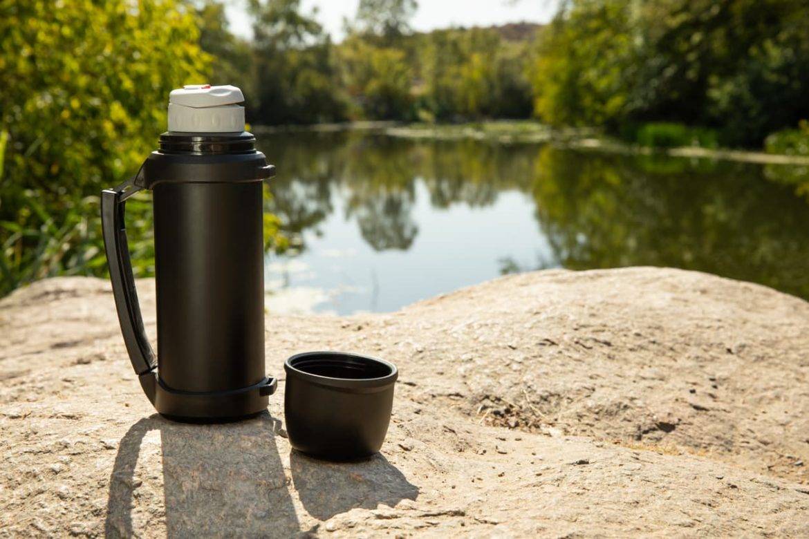 hermos with delicious hot tea or coffee on the mountain. Autumn forest background is blurred. Warming drink in a thermos against the backdrop of a lake in the forest