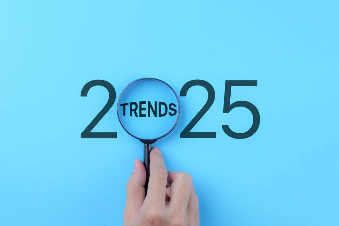 A hand holds a magnifying glass focusing on the word "Trends" within the year "2025," representing the concept of future business and marketing trends and planning.